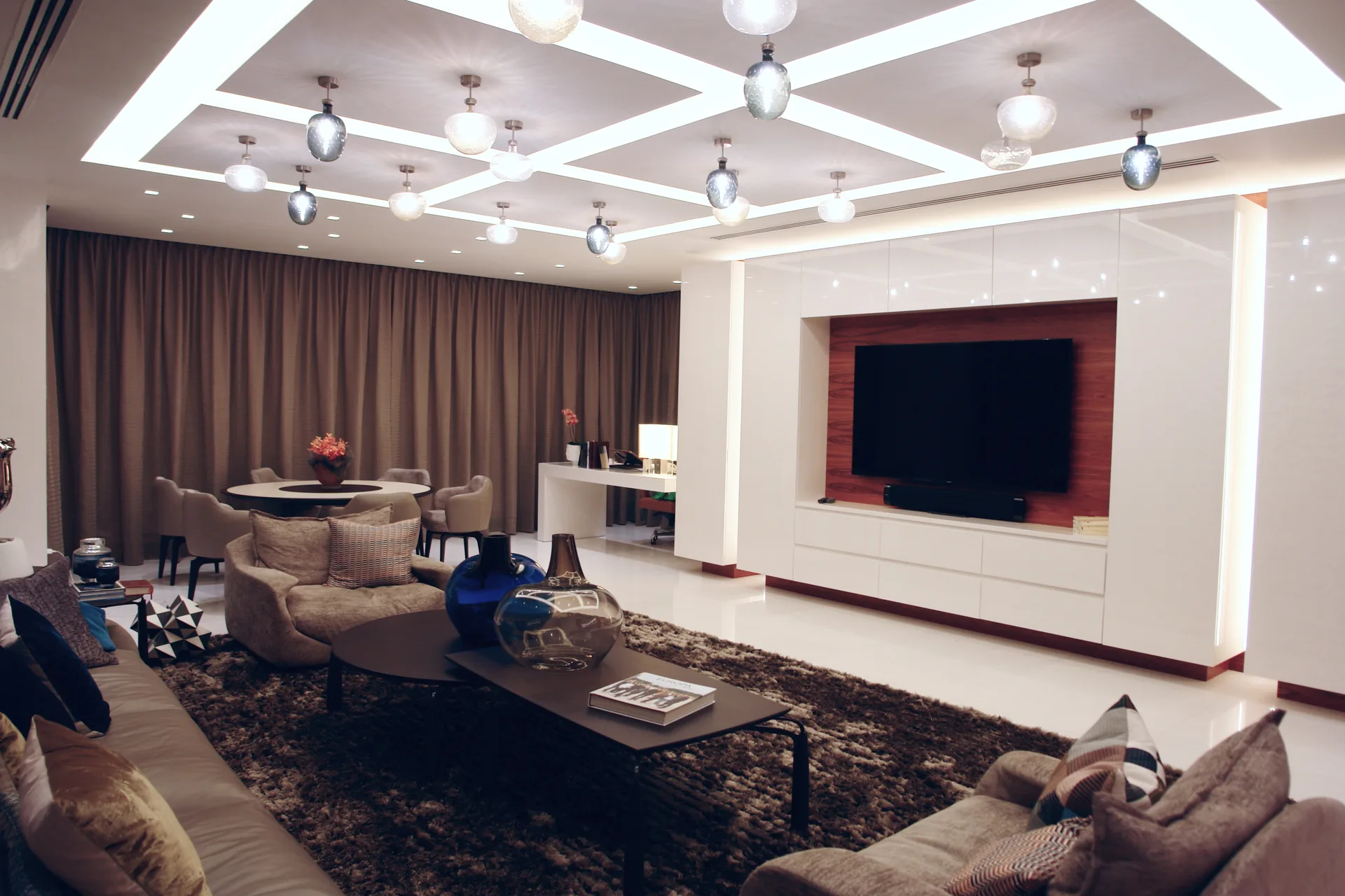 Salón con techo artesonado retroiluminado, lámparas de cristal y mueble de televisión lacado